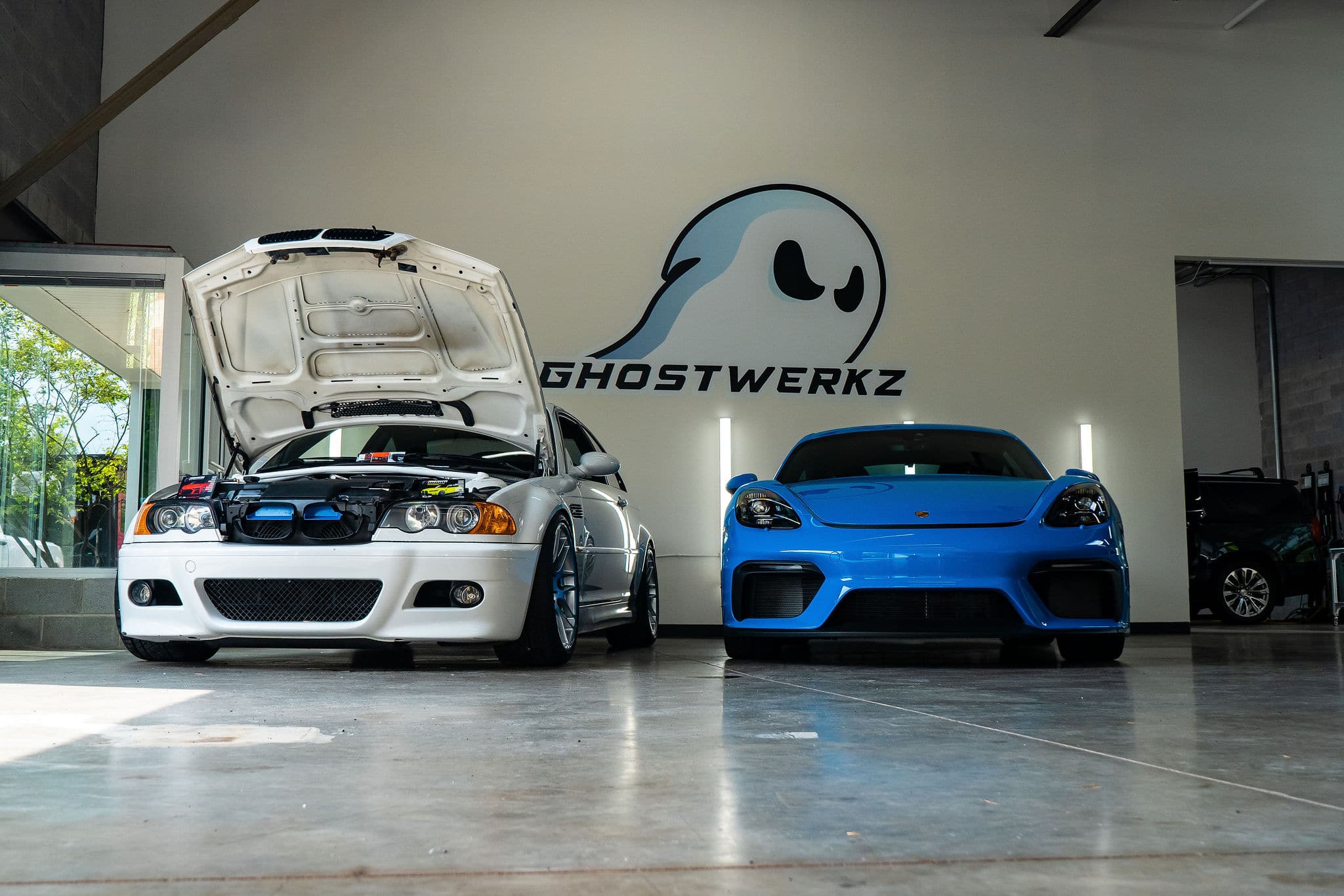White BMW with its hood open and a blue Porsche parked side-by-side inside the Ghostwerkz automotive shop.